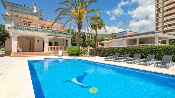 A swimming pool with lounge chairs, a building with a balcony, and palm trees.
