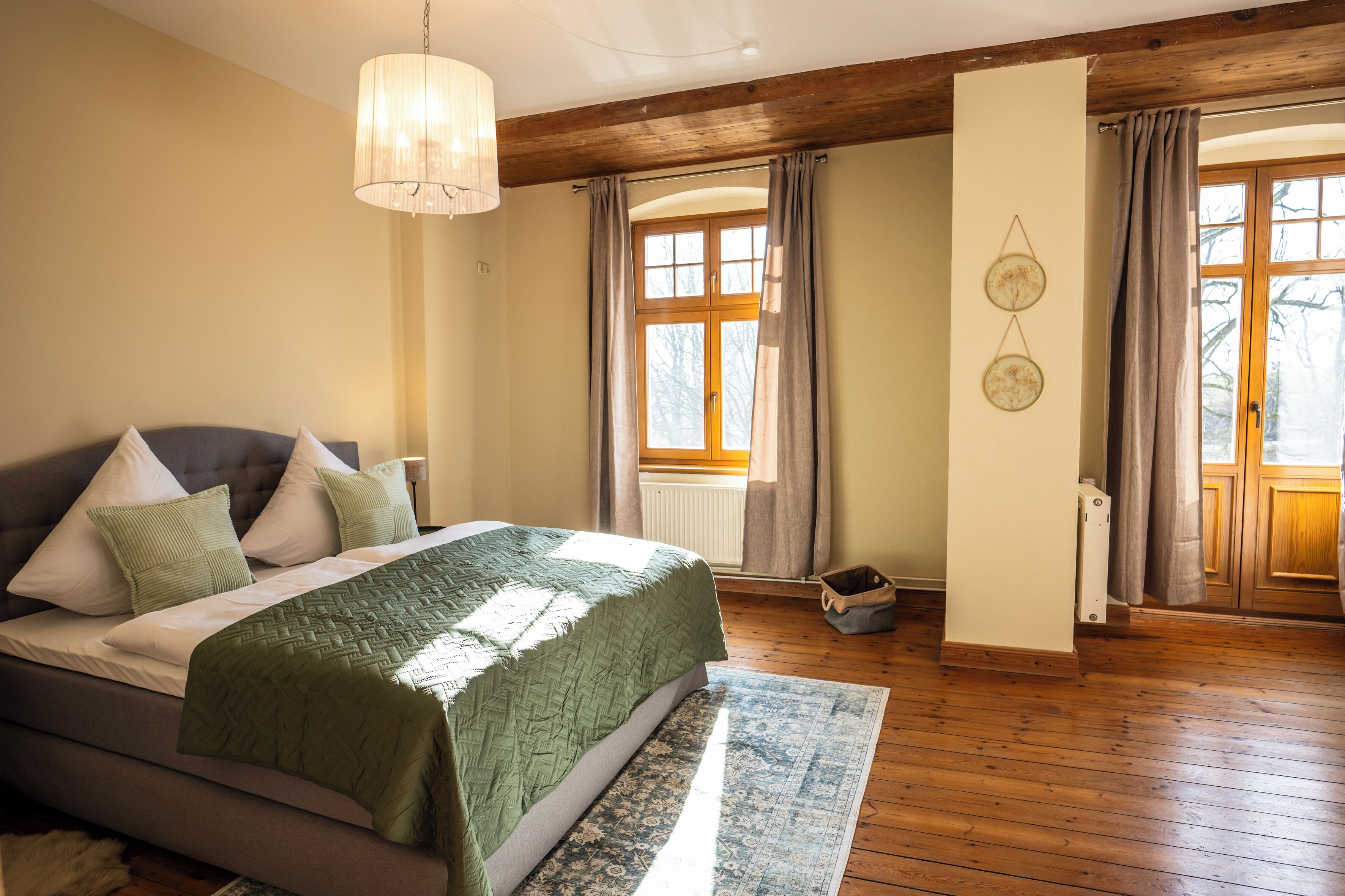 A bedroom with a bed, wooden floor, a hanging light fixture, and a window with curtains.