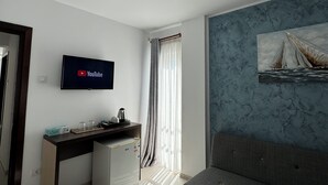 A modern room with a flat-screen TV mounted on the wall, a desk with a kettle, a sofa, and a painting of sailboats on the wall.