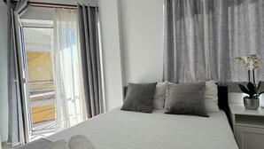 A neatly made bed with grey linens, a bedside table with a plant, and a window with curtains.