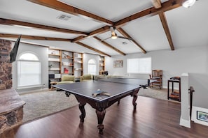 Una habitación amplia con mesa de ping pong, vigas de madera, chimenea de piedra y una estantería llena de libros y objetos decorativos.