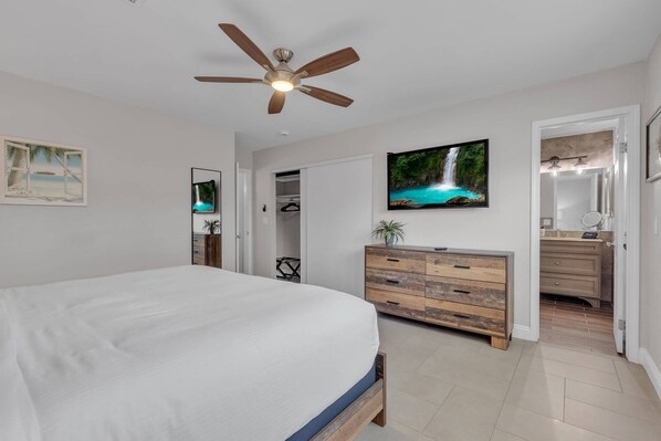A bedroom with a large bed, a ceiling fan, a TV, a wooden dresser, and a bathroom visible through an open door.