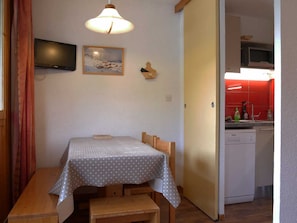 Une petite pièce comprenant un lit, une table, un téléviseur fixé au mur et un coin cuisine avec des carreaux rouges.