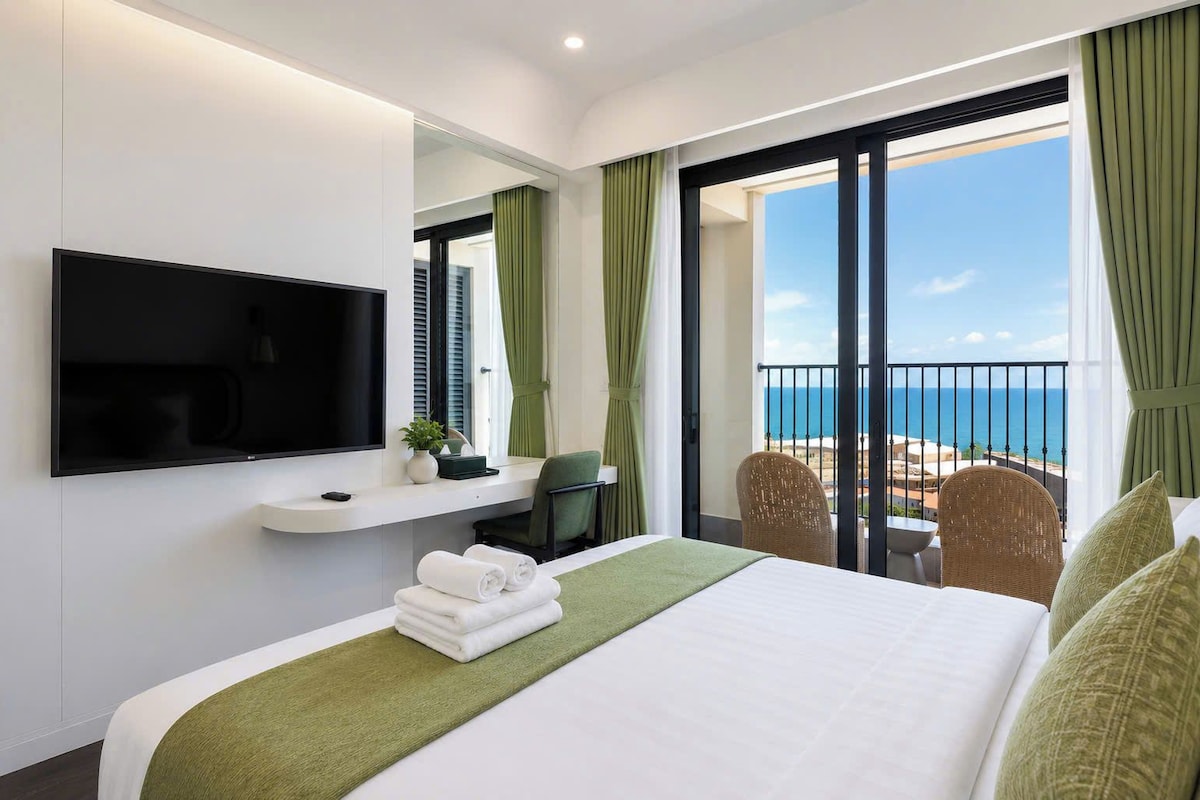 A hotel room with a bed, a TV, a small table with a plant, a chair, and a balcony with a view of the sea.
