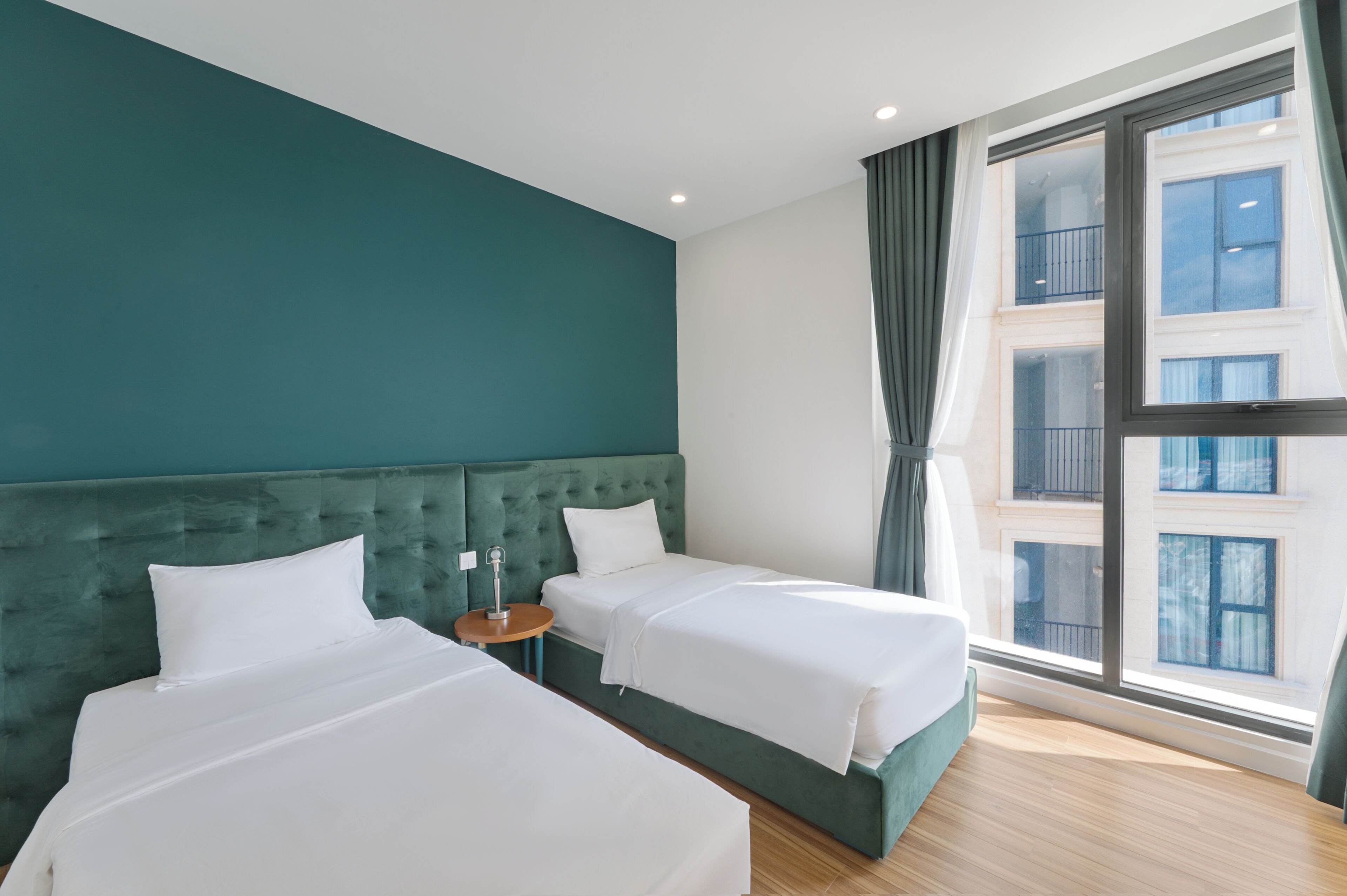 A modern hotel room with two beds, a small round table, and a large window with curtains.