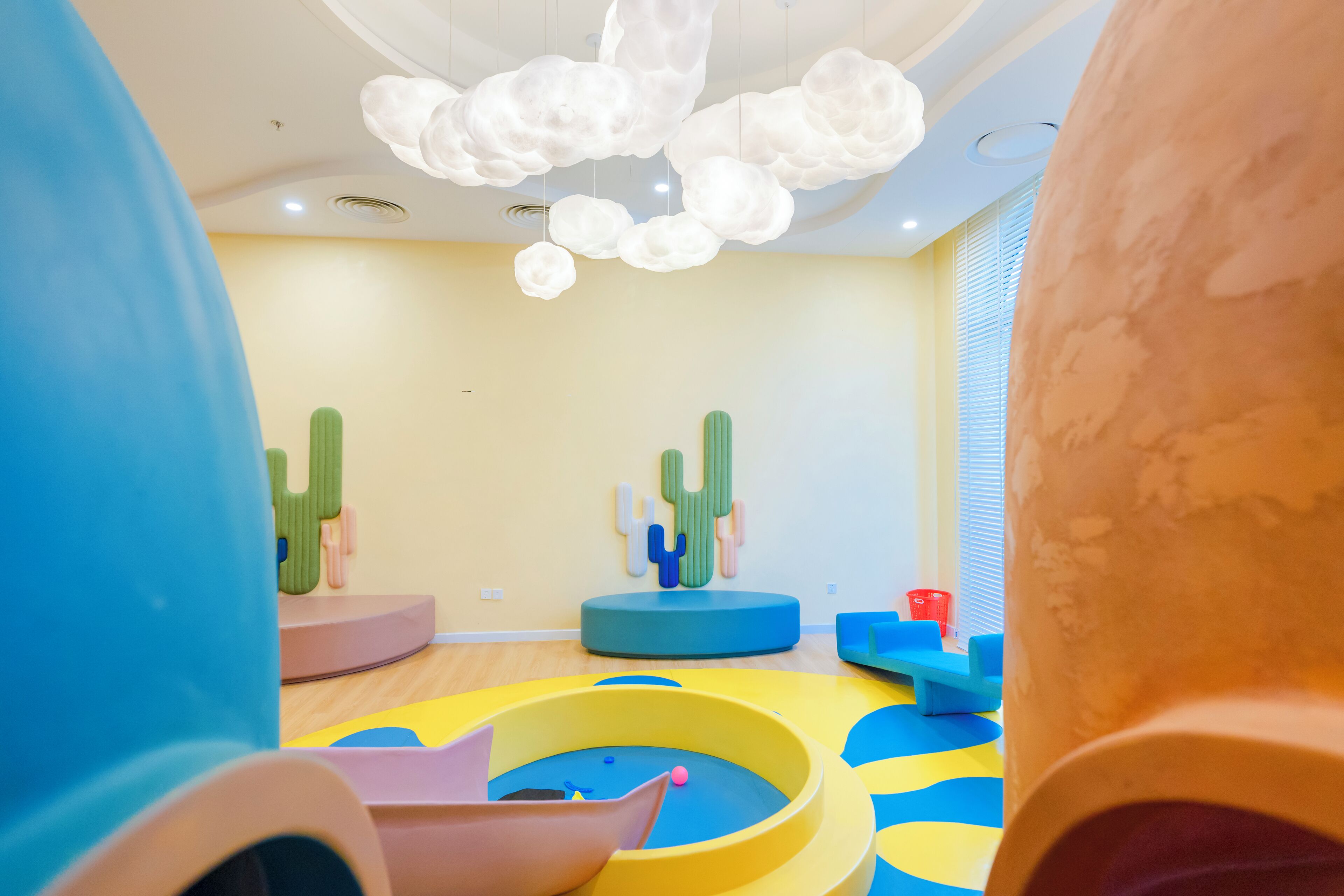 Uma área de recreação infantil com estruturas coloridas, teto com luzes em formato de nuvens e janela com persianas.