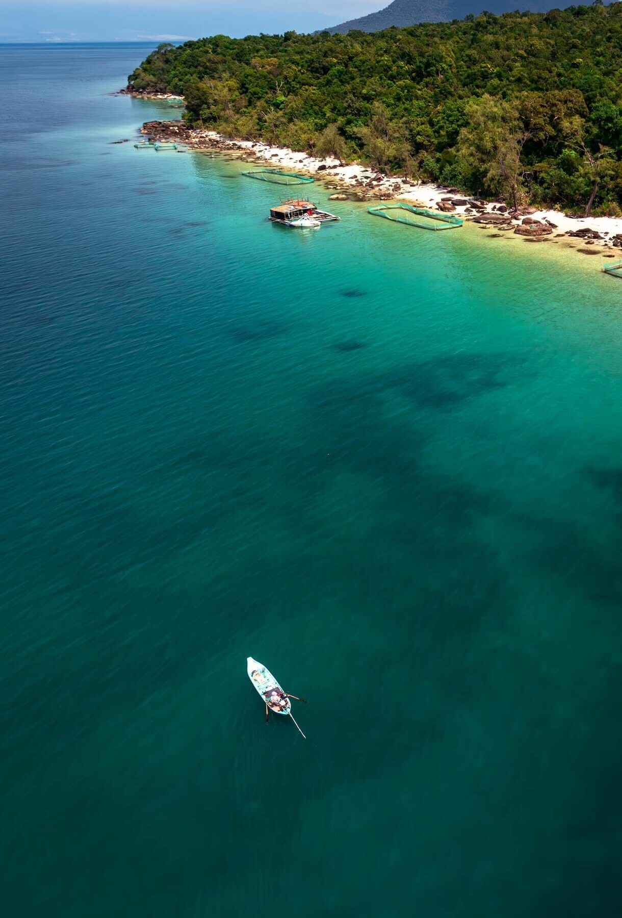 An aerial view of a coastal area with clear turquoise waters, a small boat, and a beach with dense vegetation.