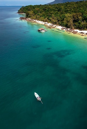An aerial view of a coastal area with clear turquoise waters, a small boat, and a beach with dense vegetation.