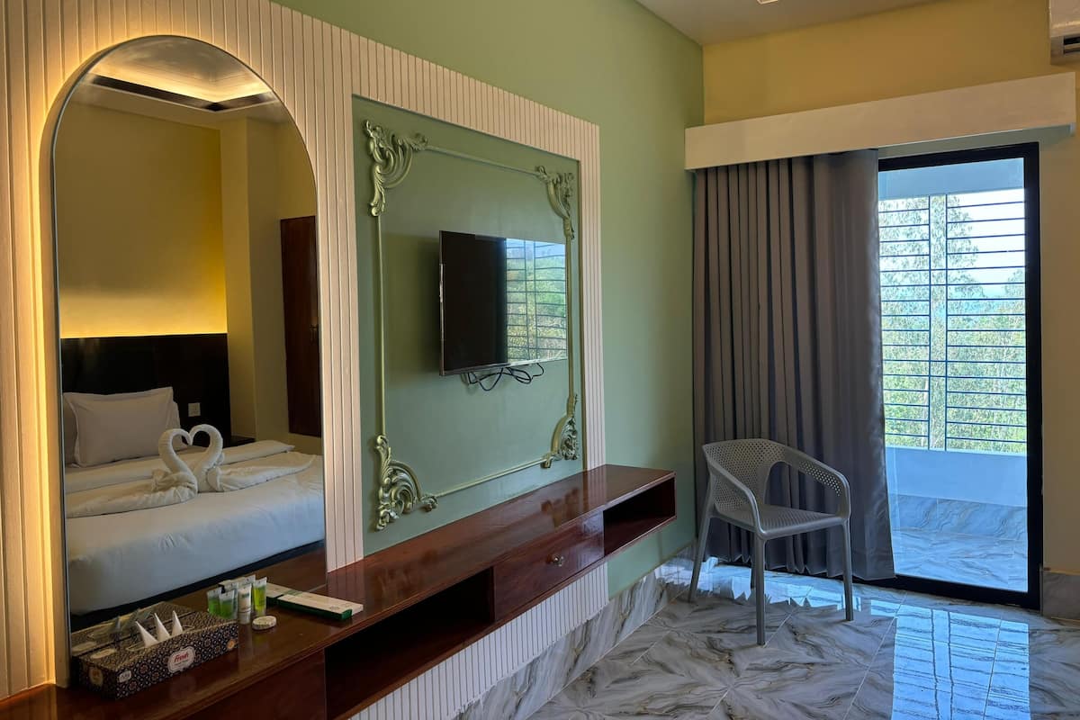 Une chambre d’hôtel comprenant un lit, un téléviseur fixé au mur, un miroir, une coiffeuse avec un miroir, une chaise et une fenêtre avec des rideaux.