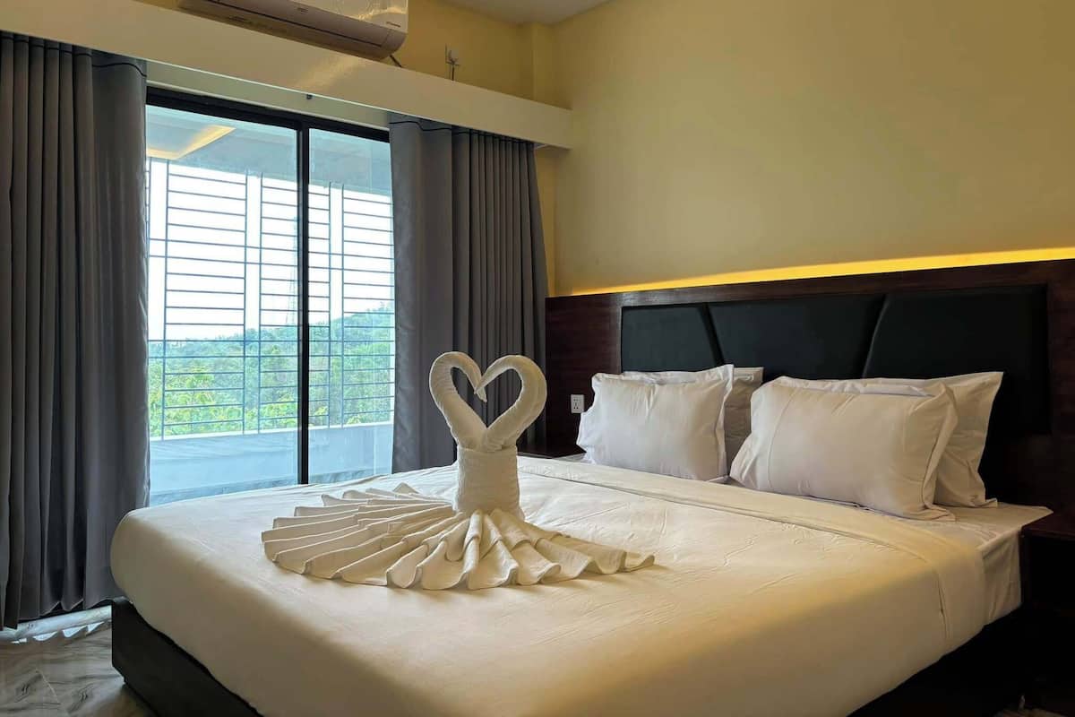 Une chambre d’hôtel avec un lit parfaitement fait, une décoration en forme de cœur faite avec des serviettes, une grande fenêtre avec des rideaux et un climatiseur fixé au plafond.