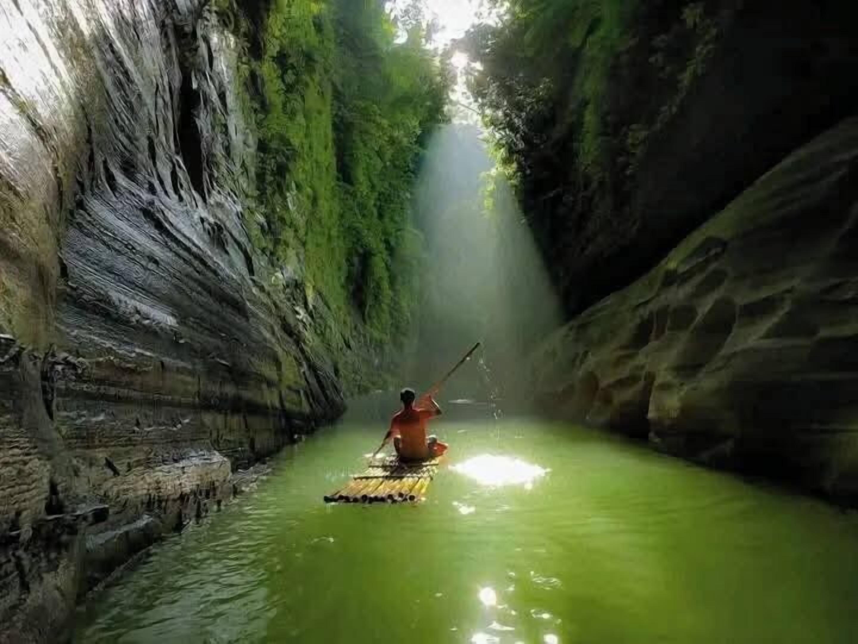 Uma pessoa remar um canoa por um canal estreito e esverdeado, ladeado por paredes rochosas.