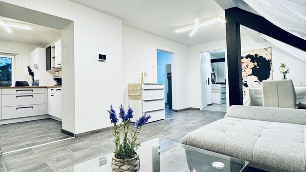 Een moderne woonkamer met een glazen salontafel, een bank en een keukenhoek met witte kasten en een blauwe deur.