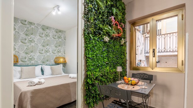 Una habitación con cama, una mesa y sillas, un ventanal y una pared verde con plantas.