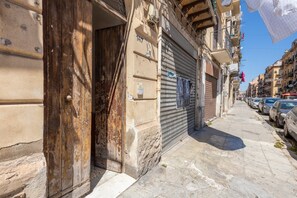 Une rue étroite, bordée de boutiques fermées, de voitures garées et d’un immeuble avec un volet métallique.