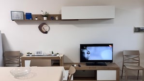 A modern living room with a dining table, chairs, a television, a shelf with decorative items, and a clock. - Sole d'Otranto, independent apartment in Otranto (punta area) 500m from the sea (Otranto)