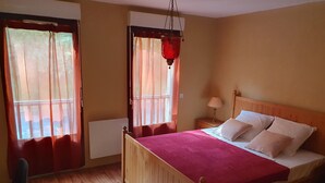 A wooden bed with a red bedspread and white pillows in a room with wooden flooring and a window with sheer curtains. - Studio in residence near Lourdes / Vallée des Gaves (Aspin-en-Lavedan)