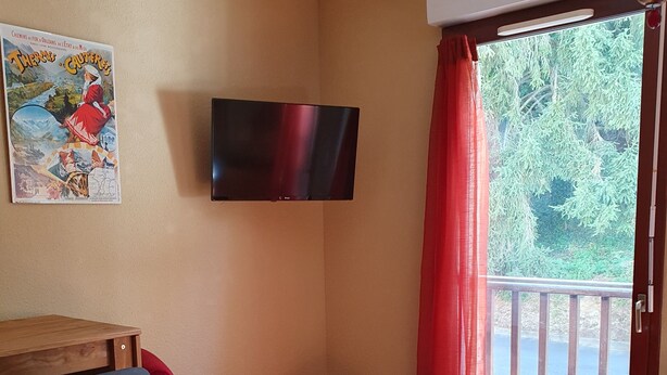A room with a wooden floor, a TV mounted on the wall, a small table with a chair, and a window with red curtains. - Studio in residence near Lourdes / Vallée des Gaves (Aspin-en-Lavedan)