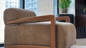 Un fauteuil moderne, doté d’un revêtement texturé et d’une structure en bois, est placé dans une pièce avec de grandes fenêtres, une plante en pot et une vue sur un bâtiment à l’extérieur.