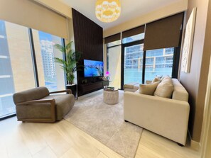 A modern living room with a large flat-screen TV, comfortable seating, a potted plant, and large windows offering a city view. - Pool View 1BR | Modern & Stylish (Dubai)