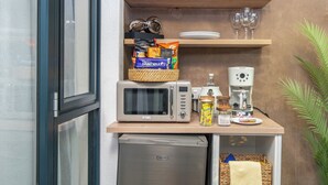 Eine Küchenzeile mit Mikrowelle, Kaffeemaschine und kleinem Kühlschrank. Darüber befinden sich Regale mit Geschirr, Gläsern und einem dekorativen Gegenstand.