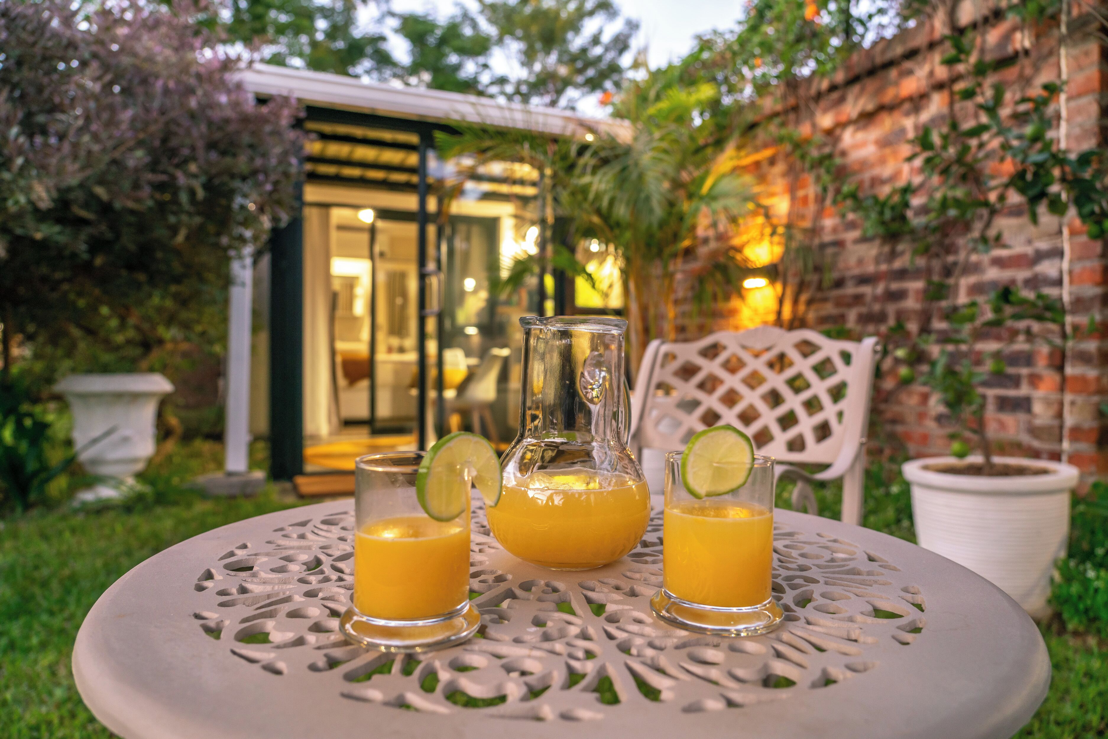 A table with three glasses of orange juice and a pitcher, set outdoors with a brick wall and greenery in the background.