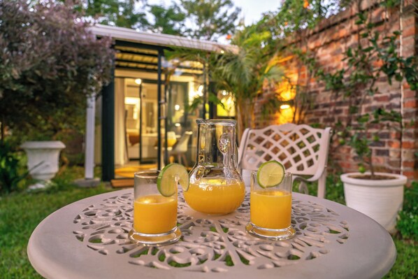 A table with three glasses of orange juice and a pitcher, set outdoors with a brick wall and greenery in the background.