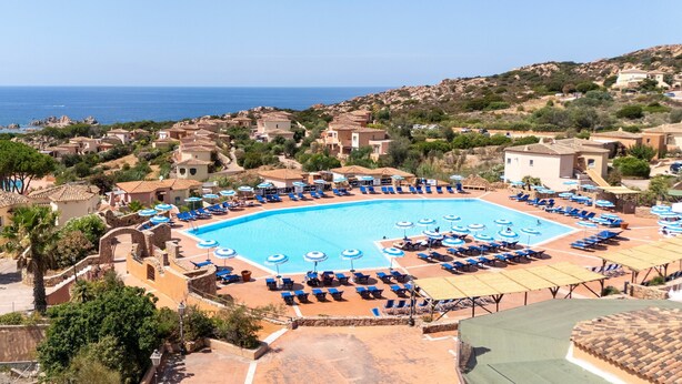 A resort with a large swimming pool surrounded by sun loungers and umbrellas, set against a backdrop of hills and the sea.