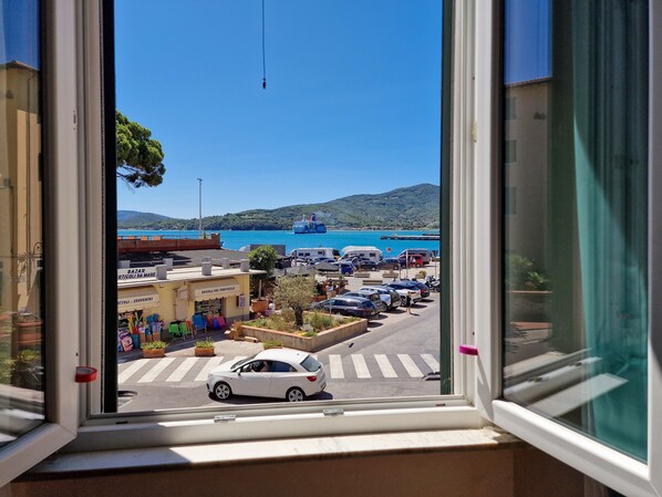 A view through a window of a coastal town with parked cars, a street crossing, and a ferry in the background.