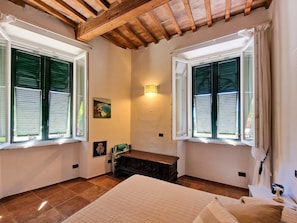 A bedroom with two windows, a wooden ceiling, a bedside table, and a bed.