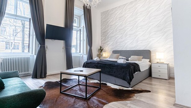 A modern bedroom with a bed, a TV mounted on the wall, a chandelier, and a view of the city. - Affordable Hotel Room with City view | Mariahilfer (Wien)