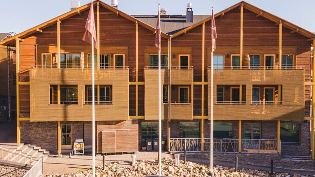 Un edificio de madera de varios pisos, con balcones, ventanas amplias y un cimiento de piedra.