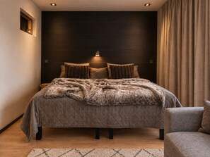 Quarto moderno com cama grande, cabeceira escura e uma cadeira lateral.