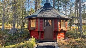 Une petite cabane en bois rouge, avec une cheminée, entourée d’une forêt.