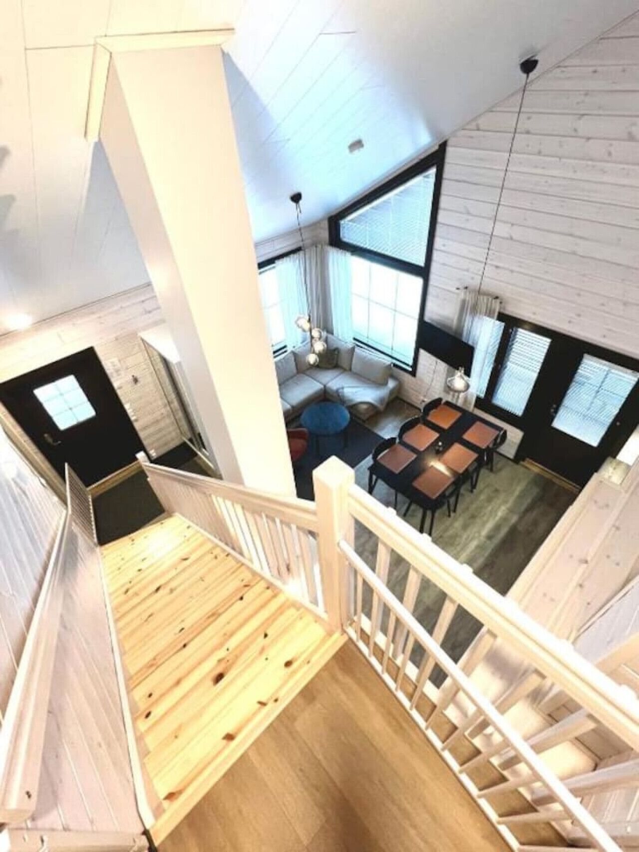 Un interior moderno con una escalera de madera, una zona de estar con sofá y mesas, y ventanas amplias que ofrecen luz natural.