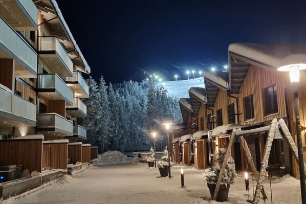 A snow-covered outdoor area with wooden buildings and a large tree in the background.
