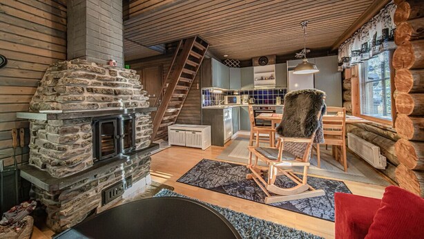 A cozy cabin interior with a stone fireplace, wooden ceiling, a dining area with a red chair, and a kitchen with a window overlooking the outside.