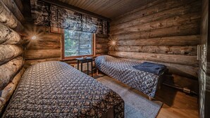 A log cabin room with two beds, a window with blinds, and a small table.