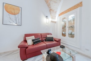 Un canapé en cuir rouge agrémenté de coussins décoratifs, une table basse en verre, un luminaire fixé au mur et une grande fenêtre donnant sur un mur de briques.