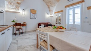 Un coin repas avec une table dressée pour deux personnes, un espace cuisine avec un comptoir et des placards, et un espace de vie avec un canapé et une plante.