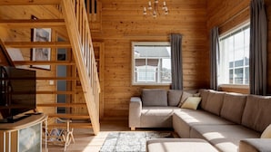 Interno di una baita in legno con un divano, una televisione, una scala e una finestra con tende.