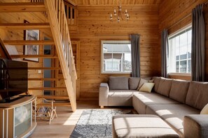 Interior de una cabaña de madera con sofá, televisor, escalera y ventana con cortinas.