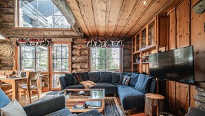 Ein rustikaler Innenraum einer Blockhütte mit einem großen Ecksofa, einem Flachbildfernseher und einer Essecke mit Holztischen und -stühlen.