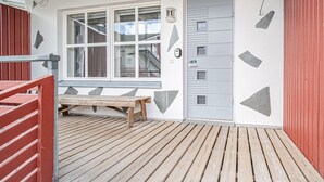 Eine Holzterrasse mit einer Bank, einer Tür mit einer Nummernschild und einem Fenster mit Blick auf ein Gebäude.
