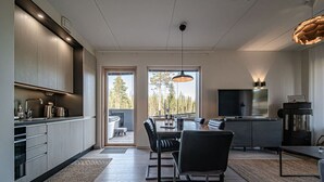 Una cocina moderna con comedor, televisor y vistas a árboles por las ventanas.