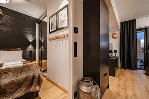 A modern hotel room with a wooden floor, a bed with a brown cover, a wooden bench, a small table with a lamp, and a large window with black curtains.
