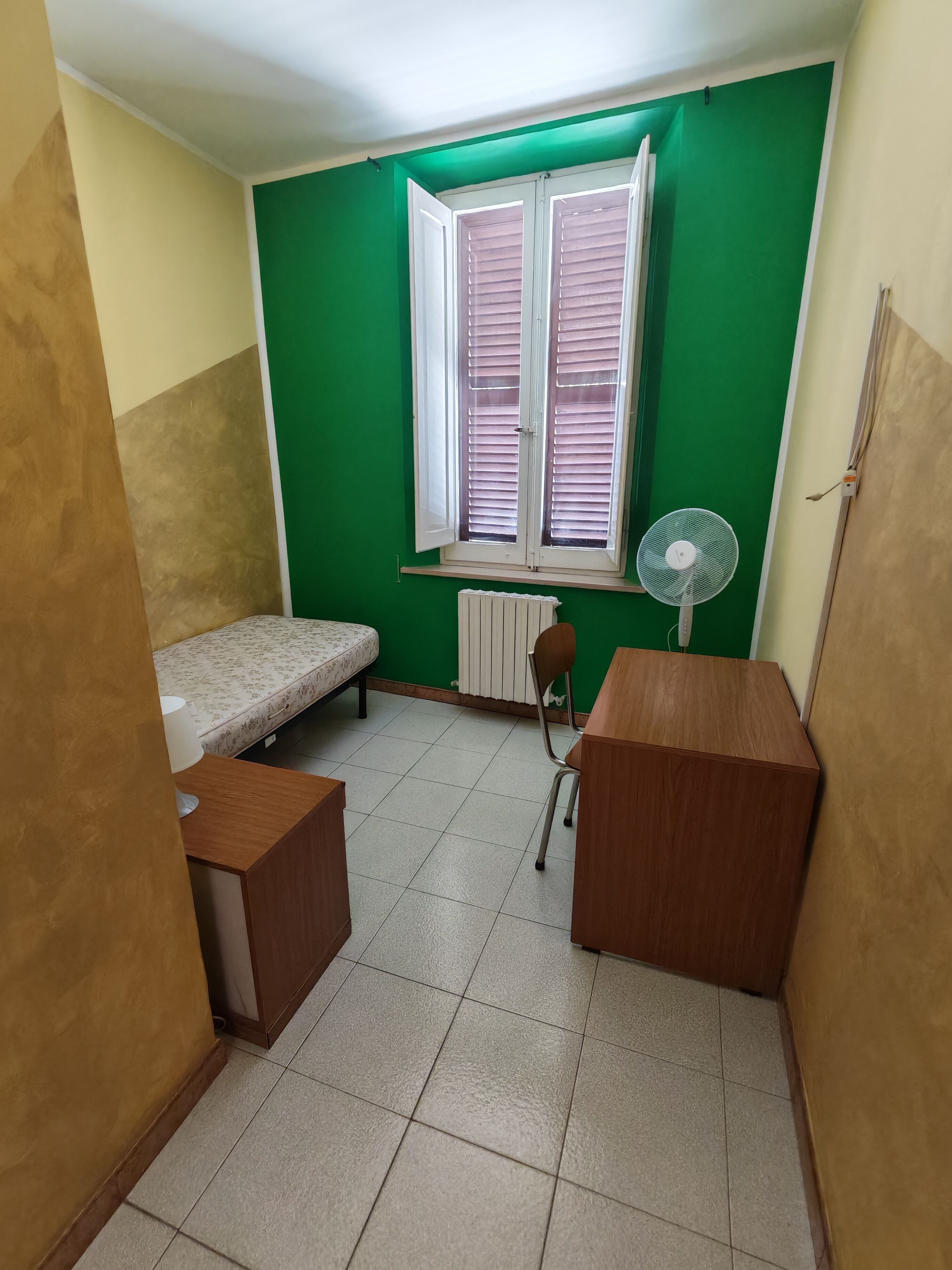 A small, compact room with a single bed, a desk, a chair, and a radiator.