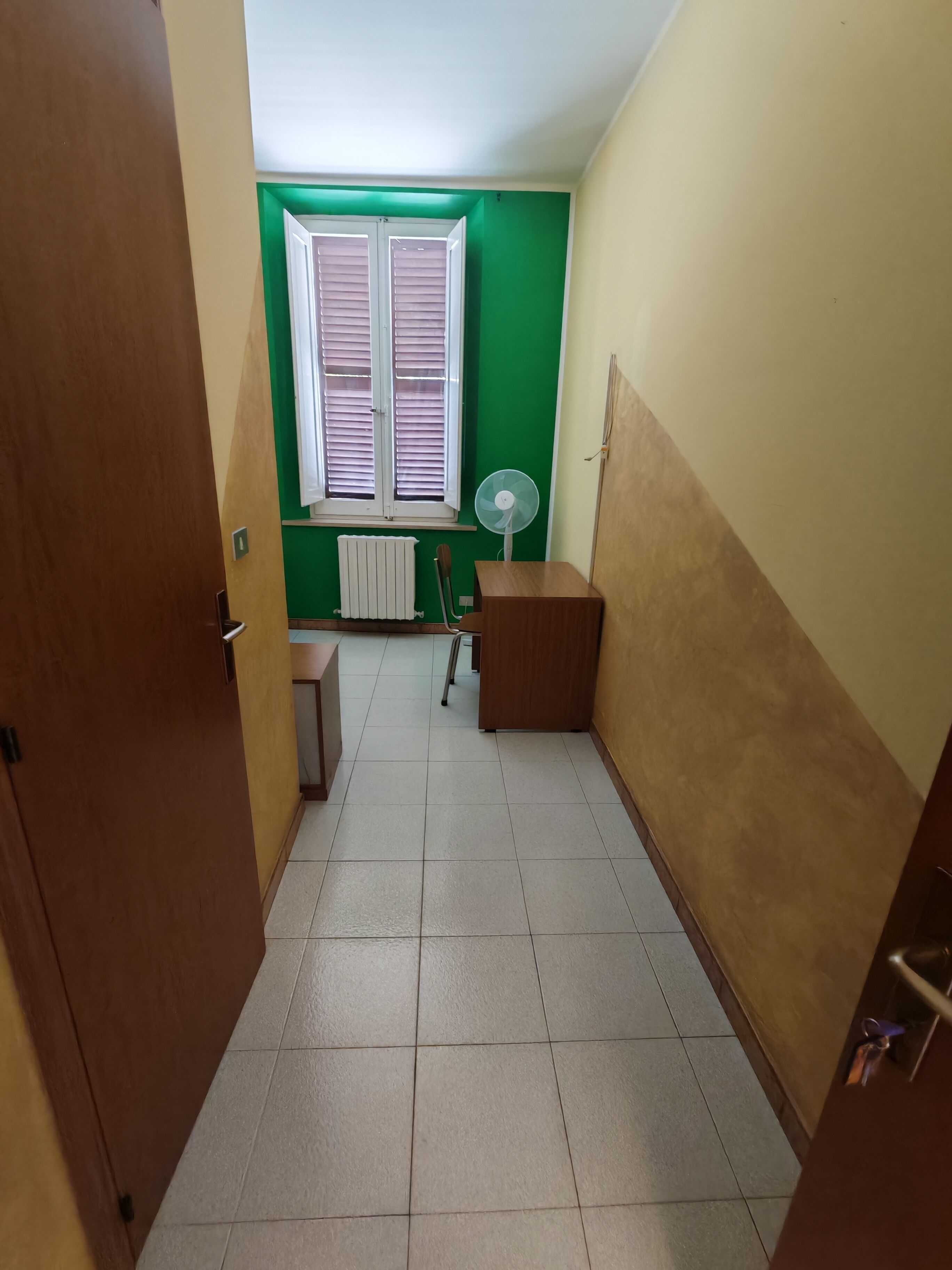 A narrow hallway with a wooden door on the left, a radiator, a desk, and a window with blinds.
