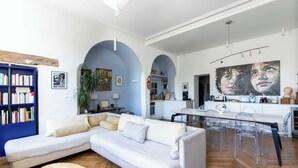 A modern living room with a white sectional sofa, a glass dining table with chairs, a bookshelf, and a kitchen area in the background. - On a southern quay of the Île Saint Louis. Like a house. Close to Notre-Dame. (Paris)