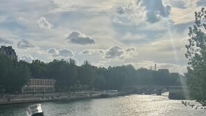 A hand holding a bottle and a glass near a river with buildings in the background. - On a southern quay of the Île Saint Louis. Like a house. Close to Notre-Dame. (Paris)