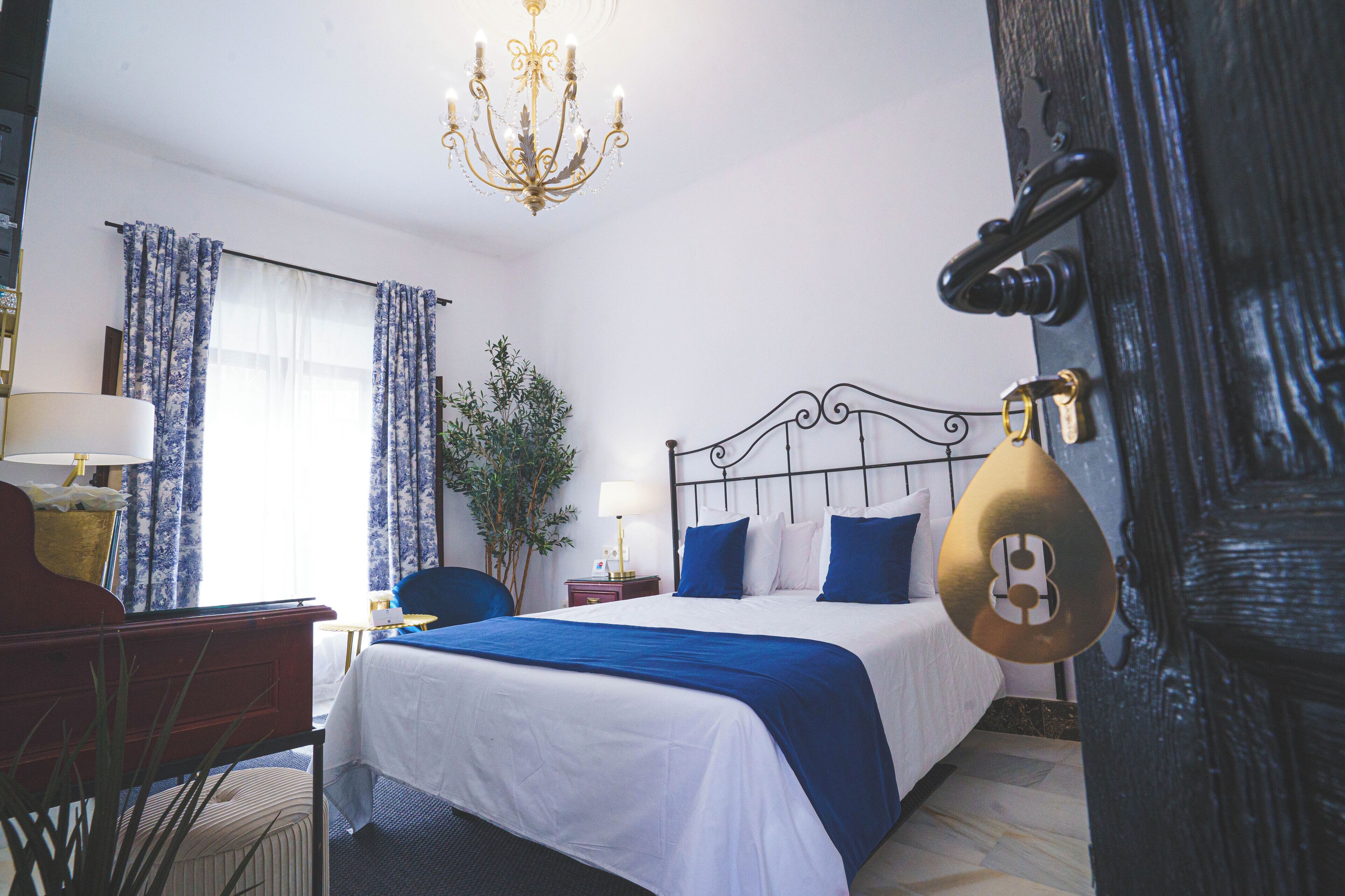 Une chambre d’hôtel avec un lit, un bureau, une chaise, une lampe, un lustre et une porte avec une serrure.